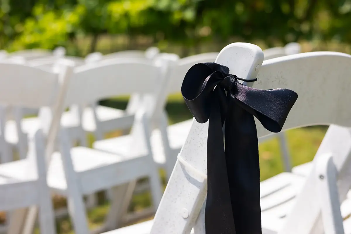 A black satin bow tied to the back of a white folding chair at an outdoor wedding ceremony