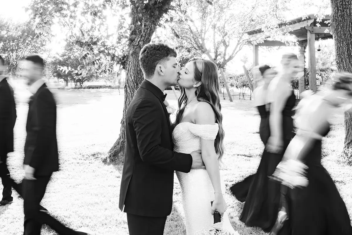 A black and white photo of a bride and groom sharing a kiss in an outdoor setting, with blurred figures of the wedding party moving around them.