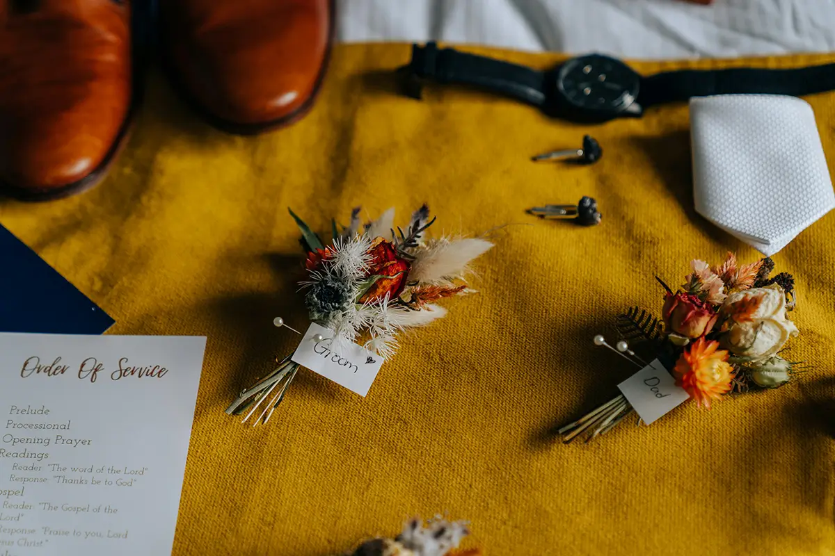 Groom's wedding accessories with autumn boutonnières on a mustard fabric backdrop.