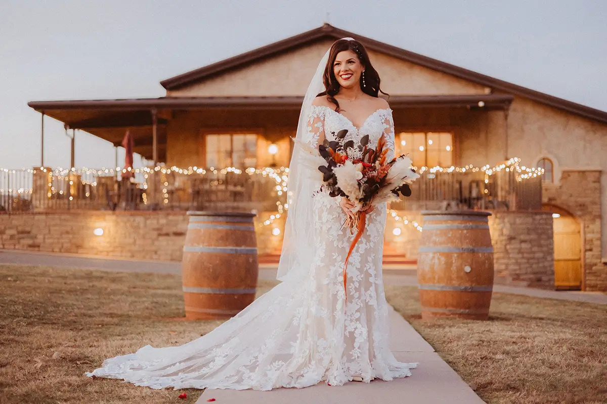 A bride in a lace wedding dress stands on a pathway holding a bouquet, with a rustic venue and string lights in the background.