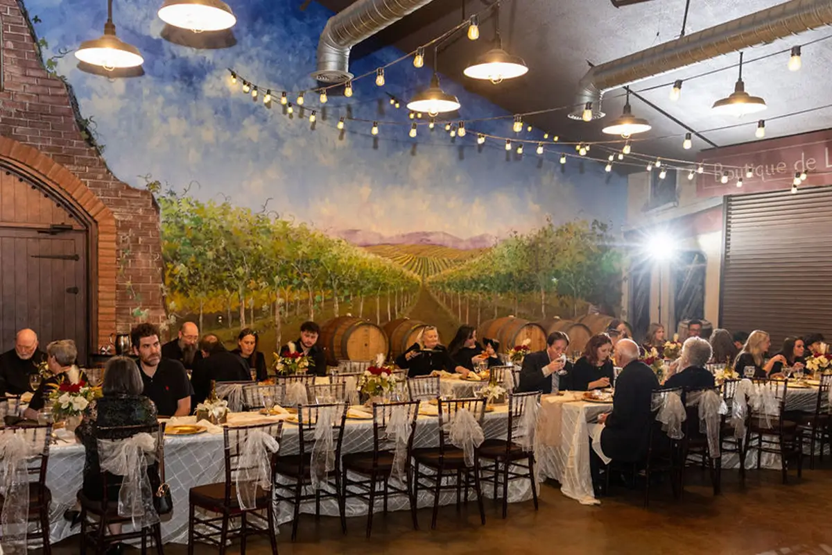 Guests seated at long tables for a banquet inside a rustic event space with a vineyard wall mural and hanging string lights.